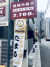 A vertical banner with a cartoon bear face and the word 'Ponta' displayed prominently. Below the bear face, more text is visible. Above, there is a maroon sign indicating pricing information in Japanese. The scene is set on a city street with tall buildings and electrical wires in the background.