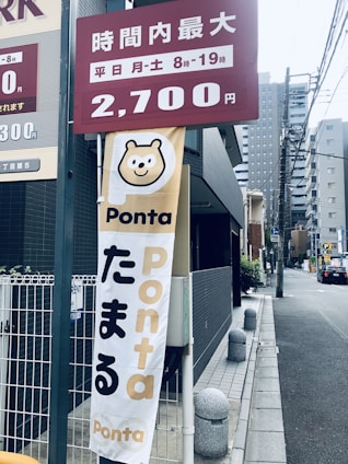 A vertical banner with a cartoon bear face and the word 'Ponta' displayed prominently. Below the bear face, more text is visible. Above, there is a maroon sign indicating pricing information in Japanese. The scene is set on a city street with tall buildings and electrical wires in the background.