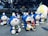 A collection of large, colorful statues featuring a cartoon character with a blue body, white face, and red nose and bow tie. Each statue holds or is surrounded by various smaller figures and objects. The setting appears to be an outdoor public space with trees in the background and people walking past.