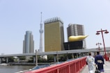 A red bridge leads across a river towards a cityscape featuring iconic modern architecture, including a tall tower and distinctive buildings with unique designs. Two people wearing traditional clothing walk along the bridge under clear skies.