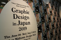 A wall displays promotional material for the Tokyo Midtown Design Hub's 80th Exhibition focused on Graphic Design in Japan 2019. The text is printed on large white ovals, with one in the foreground and others receding into the background. Numerous colorful, abstract designs are part of the artwork surrounding the text.