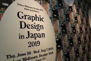 A wall displays promotional material for the Tokyo Midtown Design Hub's 80th Exhibition focused on Graphic Design in Japan 2019. The text is printed on large white ovals, with one in the foreground and others receding into the background. Numerous colorful, abstract designs are part of the artwork surrounding the text.