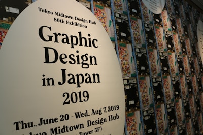 A wall displays promotional material for the Tokyo Midtown Design Hub's 80th Exhibition focused on Graphic Design in Japan 2019. The text is printed on large white ovals, with one in the foreground and others receding into the background. Numerous colorful, abstract designs are part of the artwork surrounding the text.