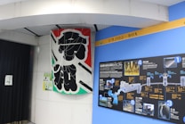 A large banner with bold black Japanese characters is displayed next to a blue wall featuring a detailed informational board. The board contains diagrams and images related to water technology or infrastructure, labeled with numbers and Japanese text. There are various colors including black, blue, and white on the board.