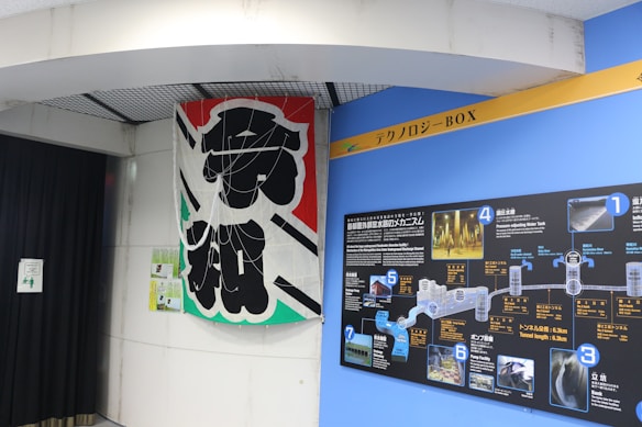 A large banner with bold black Japanese characters is displayed next to a blue wall featuring a detailed informational board. The board contains diagrams and images related to water technology or infrastructure, labeled with numbers and Japanese text. There are various colors including black, blue, and white on the board.