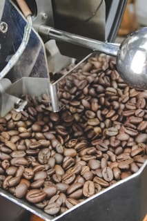 A close-up view of freshly roasted coffee beans inside a metal roasting machine. The beans are a variety of dark brown tones, indicating a range of roasting levels. The metallic parts of the machine, including a shiny pipe and a reflective surface, can be seen among the beans.