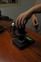 A person is preparing coffee using an AeroPress coffee maker. The setup includes an AeroPress positioned on top of a glass placed on a digital scale. A metallic kettle with a gooseneck spout is nearby, all on a wooden countertop.