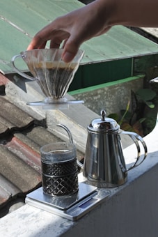 A hand is holding a glass coffee dripper filled with coffee grounds over a glass mug. Below, a shiny metal kettle is placed on a digital scale, with the scale displaying a glass filled with coffee. The setting appears to be outdoors on a rooftop or terrace with a green roof in the background.