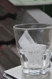 A clear glass is filled with ice cubes, placed on a metal surface with a blurred, industrial-looking background.