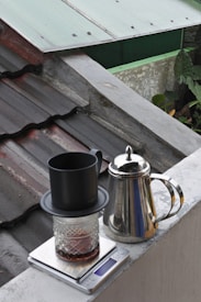 A coffee brewing setup positioned outdoors, featuring a black cup placed on top of a glass jar with a metallic dripper, all resting on a digital scale. Nearby, a shiny stainless steel kettle stands on the edge of a surface. In the background, a corrugated roof and some greenery are visible.