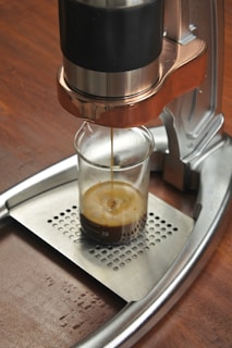 A manual espresso machine is brewing a shot of espresso into a small glass beaker placed on a stainless steel drip tray. The machine has a metallic finish with black and copper tones, and it is situated on a wooden surface.
