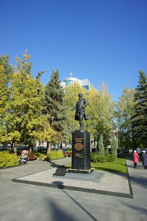 A bronze statue of a historical figure stands prominently on a pedestal surrounded by a well-maintained garden. Tall trees with lush green leaves frame the scene, and a clear blue sky stretches overhead. Several people can be seen walking or sitting nearby, and a modern building with a flag is visible in the background.