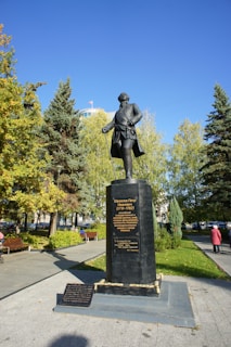 A statue of a historical figure stands prominently in a park, surrounded by lush green trees and a clear blue sky. The statue is mounted on a large pedestal with inscriptions on it. Nearby, a pathway and a person dressed in a pink jacket are visible.