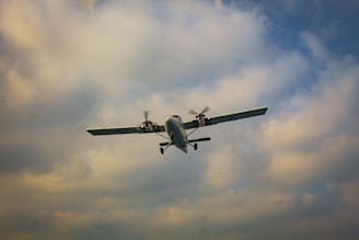 A small propeller-driven airplane is flying against a backdrop of a partly cloudy sky. The aircraft is in mid-air, with its landing gear visible and wings spread wide.