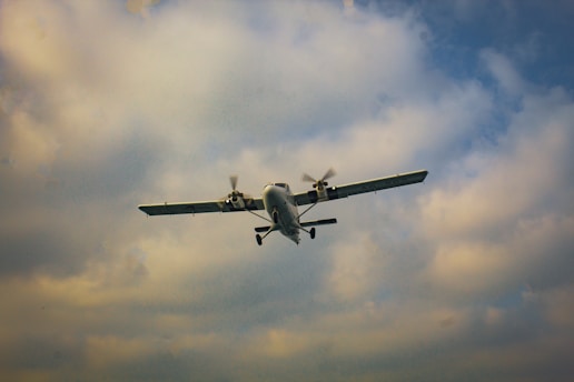 A small propeller-driven airplane is flying against a backdrop of a partly cloudy sky. The aircraft is in mid-air, with its landing gear visible and wings spread wide.