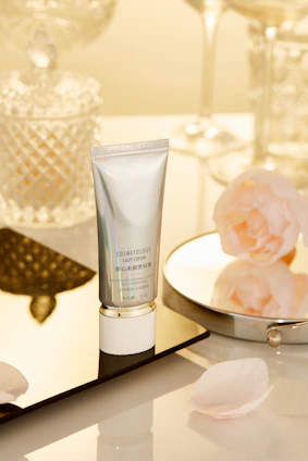 A tube labeled 'Cosmetology Lady Cream' is placed on a reflective surface. Surrounding it are decorative items such as a glass jar with a diamond pattern, a round mirror, and pink flower petals. The background features a soft, warm lighting that enhances the luxurious feel of the setting.