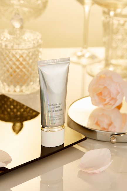 A tube labeled 'Cosmetology Lady Cream' is placed on a reflective surface. Surrounding it are decorative items such as a glass jar with a diamond pattern, a round mirror, and pink flower petals. The background features a soft, warm lighting that enhances the luxurious feel of the setting.