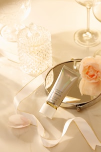 A neatly arranged composition featuring a tube of cosmetic cream placed on a circular mirror. The setup includes a delicate ribbon with gold accents, a soft pink flower, and an intricate crystal jar. The background has a warm, ambient glow created by soft lighting, with tall, elegant glassware completing the scene.