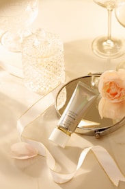 A neatly arranged composition featuring a tube of cosmetic cream placed on a circular mirror. The setup includes a delicate ribbon with gold accents, a soft pink flower, and an intricate crystal jar. The background has a warm, ambient glow created by soft lighting, with tall, elegant glassware completing the scene.