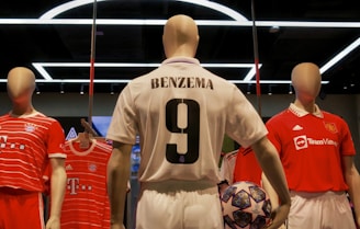 Three mannequins are dressed in football jerseys displayed in a sports store. The center mannequin wears a white jersey with 'Benzema' and the number 9 on the back, holding a soccer ball with a complex design. On the left and right, mannequins are wearing red jerseys from different football teams.