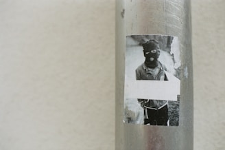 A worn, black-and-white sticker is attached to a metallic pole. The sticker features an image of a person wearing a balaclava, obscuring their face, dressed in casual wear. The background appears slightly faded, and part of the sticker is torn.