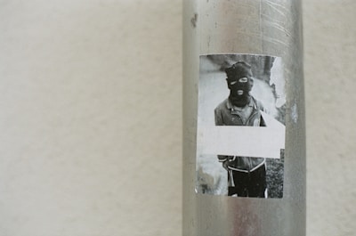 A worn, black-and-white sticker is attached to a metallic pole. The sticker features an image of a person wearing a balaclava, obscuring their face, dressed in casual wear. The background appears slightly faded, and part of the sticker is torn.