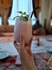 A hand holding a chilled pink smoothie topped with mint leaves and a cherry. The background shows a dining area with soft focus, featuring a table with plates and a patterned tablecloth.