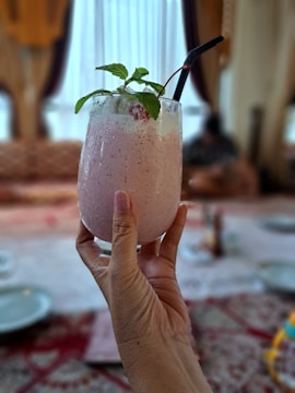 A hand holding a chilled pink smoothie topped with mint leaves and a cherry. The background shows a dining area with soft focus, featuring a table with plates and a patterned tablecloth.