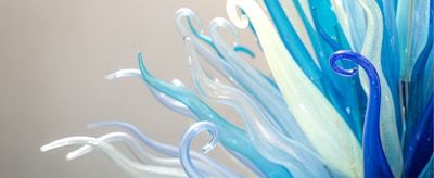 Delicate and intricately shaped glass pieces curl and twist upwards, showcasing a blend of blue and clear translucent hues. The structures resemble sea creatures or organic forms, with a smooth and glossy finish that reflects the light.