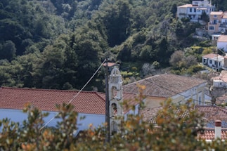 A small village with white buildings topped with red-tiled roofs is nestled in a lush, green, hilly forested area. A bell tower rises prominently in the center, featuring detailed architecture. Overhead cables are visible, crossing through the frame.
