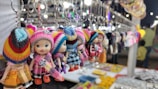 A row of colorful doll keychains with knitted hats and dresses hangs in a market stall. The focus is on the foreground dolls, with blurred market items and lights creating a vibrant background. Various items are laid out on the stall table below.