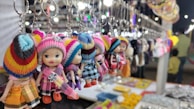 A row of colorful doll keychains with knitted hats and dresses hangs in a market stall. The focus is on the foreground dolls, with blurred market items and lights creating a vibrant background. Various items are laid out on the stall table below.