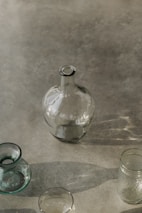 A collection of glass containers, including a large round bottle, a small vase, and a couple of jars, is arranged on a smooth, reflective surface. The lighting creates soft shadows and highlights the transparency and subtle reflections of the glass.