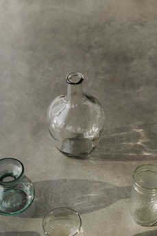 A collection of glass containers, including a large round bottle, a small vase, and a couple of jars, is arranged on a smooth, reflective surface. The lighting creates soft shadows and highlights the transparency and subtle reflections of the glass.
