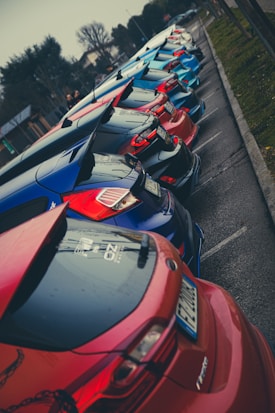 A lineup of colorful hatchback cars parked closely together along a street. The cars have distinctive rear spoilers and a sporty look. The overcast sky and wet pavement suggest a recent rain.