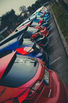 A lineup of colorful hatchback cars parked closely together along a street. The cars have distinctive rear spoilers and a sporty look. The overcast sky and wet pavement suggest a recent rain.