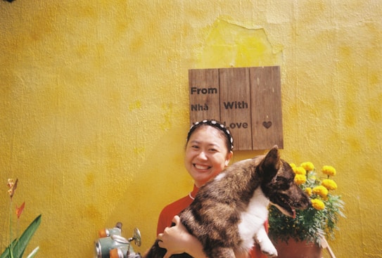 A person smiling while holding a dog against a bright yellow wall with a sign that reads 'From Nh&atilde; With Love'. Marigold flowers are in a pot beside them.