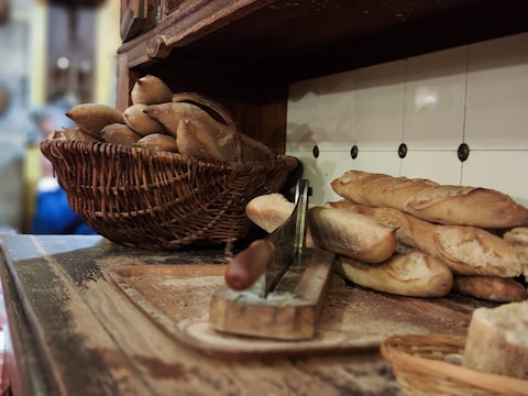 A wooden shelf holds a wicker basket filled with freshly baked baguettes. Nearby, more baguettes are arranged on a rustic wooden surface featuring a slicing board. The background consists of a tiled wall, contributing to a vintage, cozy ambiance.