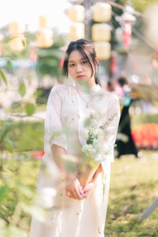 A young person wearing a light-colored, floral-patterned dress stands outdoors holding a bouquet of flowers. The background features blurred greenery and several yellow hanging lanterns.