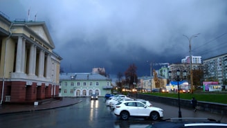 A cityscape featuring a majestic building with columns and flags, possibly a government or cultural institution, on the left. The sky is filled with dark storm clouds, creating a dramatic backdrop. Several parked cars are visible in the foreground, with a person walking near them. A lamppost and modern buildings are visible in the distance, along with some trees lining the street.