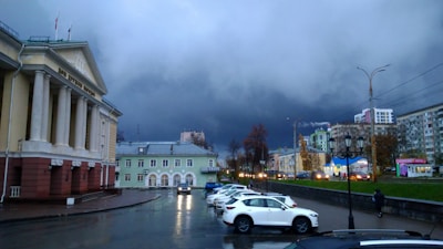 A cityscape featuring a majestic building with columns and flags, possibly a government or cultural institution, on the left. The sky is filled with dark storm clouds, creating a dramatic backdrop. Several parked cars are visible in the foreground, with a person walking near them. A lamppost and modern buildings are visible in the distance, along with some trees lining the street.