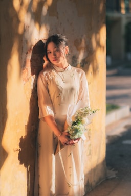 A bright, airy photo of a woman wearing a flowing orange summer dress standing against a white wall with soft shadows.