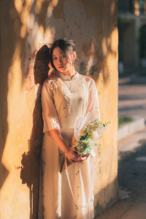 A bright, airy photo of a woman wearing a flowing orange summer dress standing against a white wall with soft shadows.