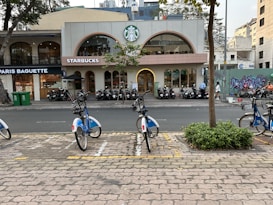 A city street scene with a row of parked bicycles in a designated bike sharing area in the foreground. Across the road, there is a Starbucks coffee shop with a distinctive logo and glass windows, next to a Paris Baguette bakery. Several motorcycles are parked in front of these establishments. The area has some greenery, with small trees and bushes. There is also graffiti visible on a wall in the background, adding to the urban atmosphere.