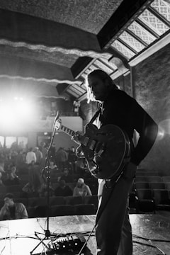 A black and white photograph captures a musician on stage holding a guitar under soft stage lighting. The musician appears pensive, looking down at the instrument. In the background, an audience is visible with some people sitting and others standing, creating a sense of anticipation. Architectural details of the venue, including patterned ceilings and walls, add to the atmosphere.
