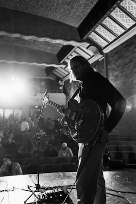 A black and white photograph captures a musician on stage holding a guitar under soft stage lighting. The musician appears pensive, looking down at the instrument. In the background, an audience is visible with some people sitting and others standing, creating a sense of anticipation. Architectural details of the venue, including patterned ceilings and walls, add to the atmosphere.