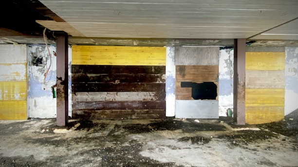 A roughly constructed wall made of various types of wooden panels, each differently colored and textured. Some parts are painted yellow, while darker brown and black wood panels are interspersed. The wall is supported by two vertical beams, and the floor underneath appears to be dirty and unfinished. The ceiling also has an unfinished look with exposed elements.