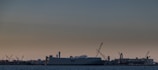 A large industrial shipyard with cranes and a massive shipping vessel is seen against the backdrop of a dim, overcast sky. The ship is anchored near structures and buildings that form part of the dockyard. The scene is set in twilight, with limited lighting affecting visibility and highlights.