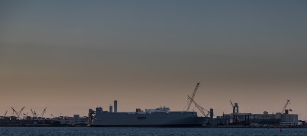 A large industrial shipyard with cranes and a massive shipping vessel is seen against the backdrop of a dim, overcast sky. The ship is anchored near structures and buildings that form part of the dockyard. The scene is set in twilight, with limited lighting affecting visibility and highlights.