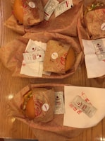 Several wrapped burgers are placed on a wooden table with packets of Dipt's tomato ketchup. Each burger is wrapped in branded paper, and white napkins are also visible beneath some of the packs.